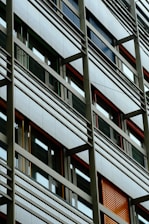 A modern building facade with a grid-like structure, featuring large windows and horizontal metal bars. The windows vary in color, with some having blue and orange louvered shutters.