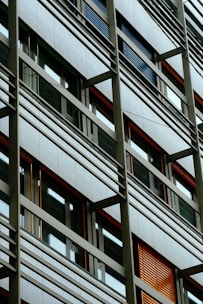 A modern building facade with a grid-like structure, featuring large windows and horizontal metal bars. The windows vary in color, with some having blue and orange louvered shutters.