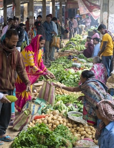 A bustling market scene with people engaging in buying and selling fresh produce. Various vegetables such as potatoes, leafy greens, and other fresh items are displayed prominently on the ground. Multiple vendors and customers are interacting, with some holding striped bags. The atmosphere is vibrant, with a mix of colorful clothing and busy activity.