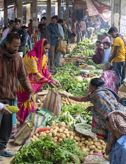 A bustling market scene with people engaging in buying and selling fresh produce. Various vegetables such as potatoes, leafy greens, and other fresh items are displayed prominently on the ground. Multiple vendors and customers are interacting, with some holding striped bags. The atmosphere is vibrant, with a mix of colorful clothing and busy activity.
