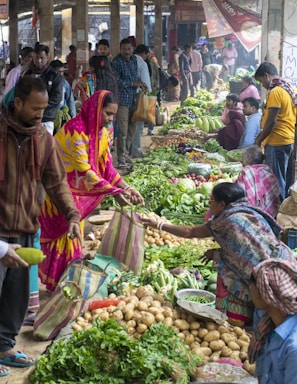 A bustling market scene with people engaging in buying and selling fresh produce. Various vegetables such as potatoes, leafy greens, and other fresh items are displayed prominently on the ground. Multiple vendors and customers are interacting, with some holding striped bags. The atmosphere is vibrant, with a mix of colorful clothing and busy activity.