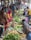A bustling market scene with people engaging in buying and selling fresh produce. Various vegetables such as potatoes, leafy greens, and other fresh items are displayed prominently on the ground. Multiple vendors and customers are interacting, with some holding striped bags. The atmosphere is vibrant, with a mix of colorful clothing and busy activity.