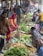 A bustling market scene with people engaging in buying and selling fresh produce. Various vegetables such as potatoes, leafy greens, and other fresh items are displayed prominently on the ground. Multiple vendors and customers are interacting, with some holding striped bags. The atmosphere is vibrant, with a mix of colorful clothing and busy activity.