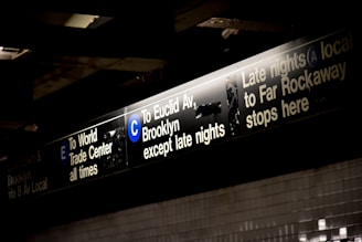 Signage for subway trains with information for lines E, C, and A, indicating directions toward the World Trade Center, Euclid Avenue Brooklyn, and Far Rockaway. The signs are black with white and blue text, illuminated from above.
