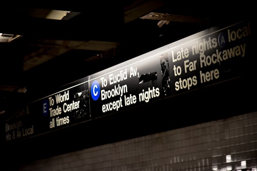 Signage for subway trains with information for lines E, C, and A, indicating directions toward the World Trade Center, Euclid Avenue Brooklyn, and Far Rockaway. The signs are black with white and blue text, illuminated from above.