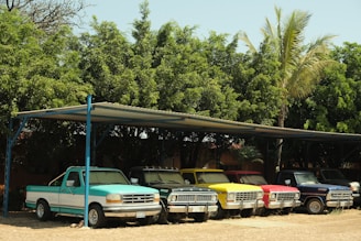 A row of colorful vintage pickup trucks is parked under a metal carport, surrounded by lush green trees and a few palm trees. The trucks are covered in dust, suggesting they haven't been used recently.