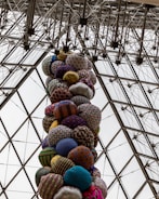 A vertical column of vibrantly patterned, round textile shapes stacked inside a geometric glass and metal structure. The patterns and colors of the textiles create a lively contrast against the structured grid of the ceiling.