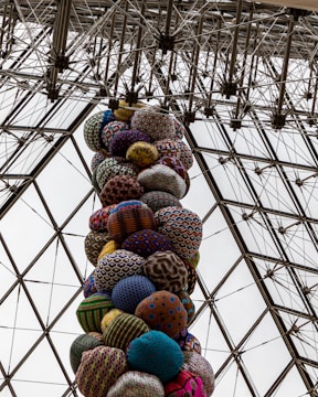 A vertical column of vibrantly patterned, round textile shapes stacked inside a geometric glass and metal structure. The patterns and colors of the textiles create a lively contrast against the structured grid of the ceiling.