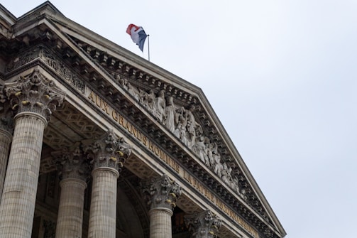 A neoclassical building with ornate Corinthian columns and a detailed pediment featuring sculpted figures. A French flag is visible on top, and an inscription reads 'AUX GRANDS HOMMES LA PATRIE RECONNAISSANTE.'
