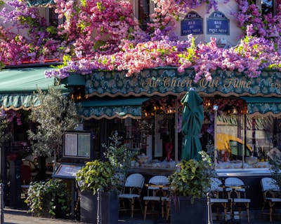 A charming caf&eacute; is adorned with vibrant, cascading flowers in shades of pink, purple, and green. Its stylish awning and outdoor seating create a cozy, inviting atmosphere. The caf&eacute; has an elegant name sign above the entrance, and the windows are lined with decorative plants, enhancing the quaint and picturesque setting.