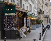A picturesque Parisian street features a charming café brasserie adorned with festive lights and floral decorations. Several tables with woven chairs are set for diners along the sidewalk. The scene is framed by classic European architecture with wrought iron balconies.