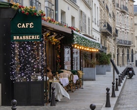 A picturesque Parisian street features a charming café brasserie adorned with festive lights and floral decorations. Several tables with woven chairs are set for diners along the sidewalk. The scene is framed by classic European architecture with wrought iron balconies.