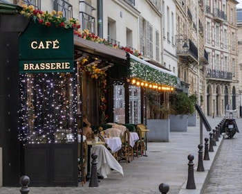 A picturesque Parisian street features a charming café brasserie adorned with festive lights and floral decorations. Several tables with woven chairs are set for diners along the sidewalk. The scene is framed by classic European architecture with wrought iron balconies.