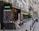 A picturesque Parisian street features a charming café brasserie adorned with festive lights and floral decorations. Several tables with woven chairs are set for diners along the sidewalk. The scene is framed by classic European architecture with wrought iron balconies.