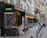A picturesque Parisian street features a charming café brasserie adorned with festive lights and floral decorations. Several tables with woven chairs are set for diners along the sidewalk. The scene is framed by classic European architecture with wrought iron balconies.