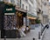 A picturesque Parisian street features a charming café brasserie adorned with festive lights and floral decorations. Several tables with woven chairs are set for diners along the sidewalk. The scene is framed by classic European architecture with wrought iron balconies.