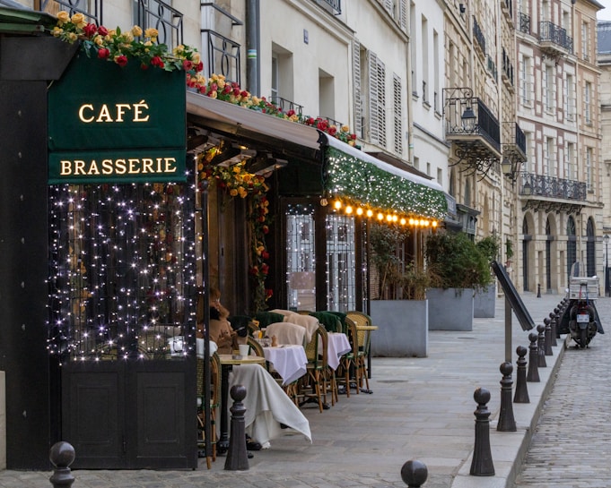 A picturesque Parisian street features a charming caf&eacute; brasserie adorned with festive lights and floral decorations. Several tables with woven chairs are set for diners along the sidewalk. The scene is framed by classic European architecture with wrought iron balconies.