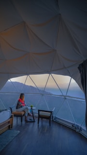 A person sits inside a dome-shaped structure with large glass panels, looking out at a mountainous landscape. The scene is indoors with wooden flooring and a cozy setup, including a chair, small table, and bed with blankets. The atmosphere appears calm and reflective.