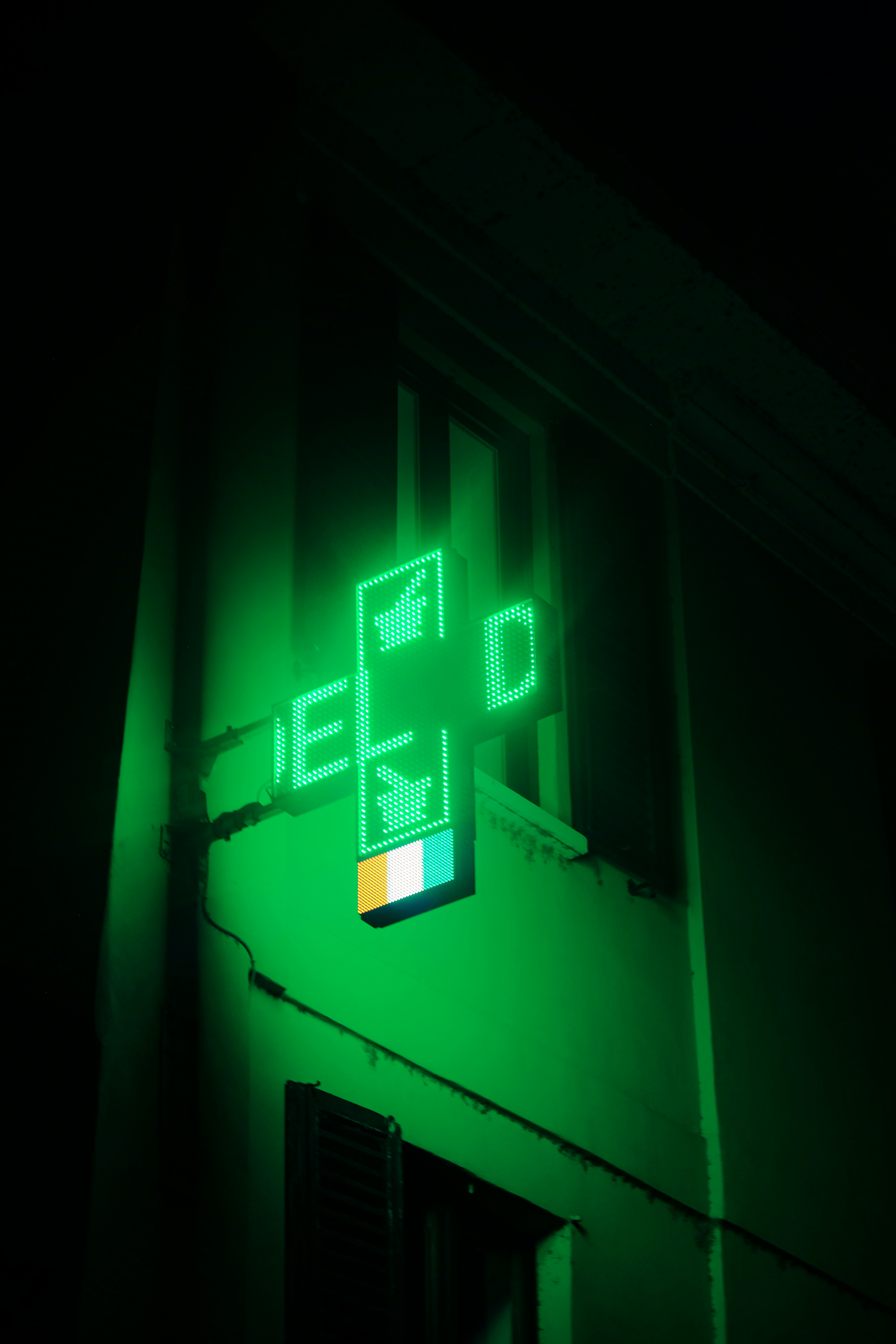 A green illuminated cross-shaped sign with digital displays, typically used to indicate a pharmacy, is affixed to the side of a building. The light from the sign casts a green hue on the surrounding area, creating a glowing effect.