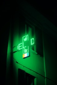 A green illuminated cross-shaped sign with digital displays, typically used to indicate a pharmacy, is affixed to the side of a building. The light from the sign casts a green hue on the surrounding area, creating a glowing effect.