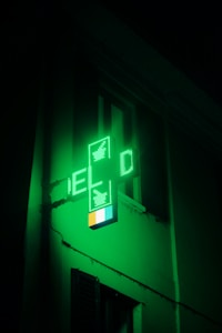 A green illuminated cross-shaped sign with digital displays, typically used to indicate a pharmacy, is affixed to the side of a building. The light from the sign casts a green hue on the surrounding area, creating a glowing effect.