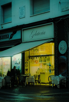 A cozy gelateria with outdoor seating, featuring small tables with chairs set up under an awning. The warm, inviting light from the shop interior contrasts with the dimly lit exterior. A person is visible near the entrance, and the shop sign is prominently displayed.
