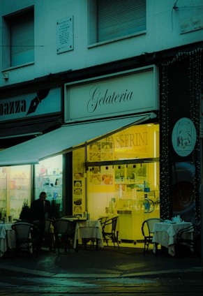 A cozy gelateria with outdoor seating, featuring small tables with chairs set up under an awning. The warm, inviting light from the shop interior contrasts with the dimly lit exterior. A person is visible near the entrance, and the shop sign is prominently displayed.