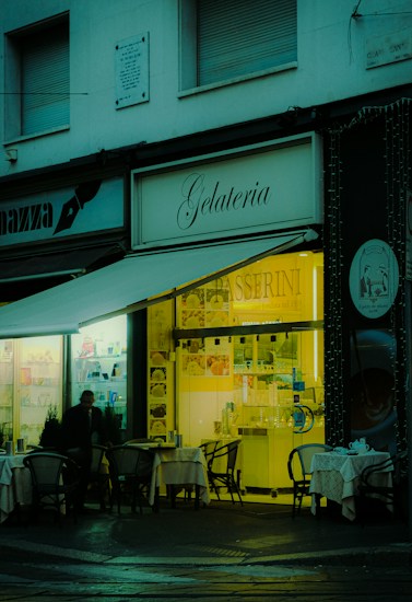 A cozy gelateria with outdoor seating, featuring small tables with chairs set up under an awning. The warm, inviting light from the shop interior contrasts with the dimly lit exterior. A person is visible near the entrance, and the shop sign is prominently displayed.