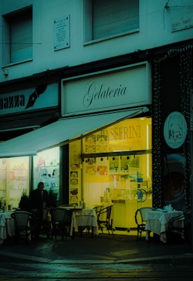 A cozy gelateria with outdoor seating, featuring small tables with chairs set up under an awning. The warm, inviting light from the shop interior contrasts with the dimly lit exterior. A person is visible near the entrance, and the shop sign is prominently displayed.