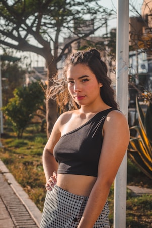 A young woman with long hair stands outdoors on a sunny day. She wears a black one-shoulder top and a checkered skirt. Her hair is styled with small braids, and she has natural makeup with a focus on her eyes, accentuated by winged eyeliner. The background includes trees, foliage, and an urban environment.