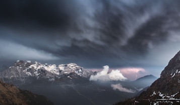 A dramatic mountain landscape under a dark and turbulent sky. Snow-covered peaks are illuminated by dim lighting, with noticeable storm clouds overhead. A bolt of lightning is visible in the distance, adding to the ominous atmosphere. Wisps of cloud hover near the mountain tops, and patches of snow are visible on the foreground hillside.