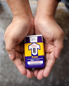 Hands holding a naloxone auto-injector device labeled Evzio, used for opioid emergencies. The device is yellow and purple with instructions for use printed on it.