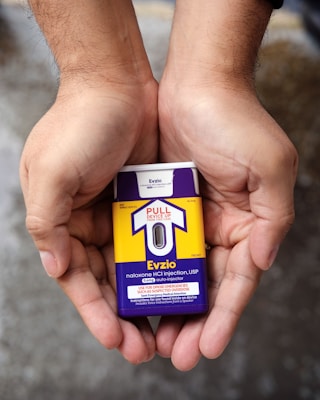 Hands holding a naloxone auto-injector device labeled Evzio, used for opioid emergencies. The device is yellow and purple with instructions for use printed on it.