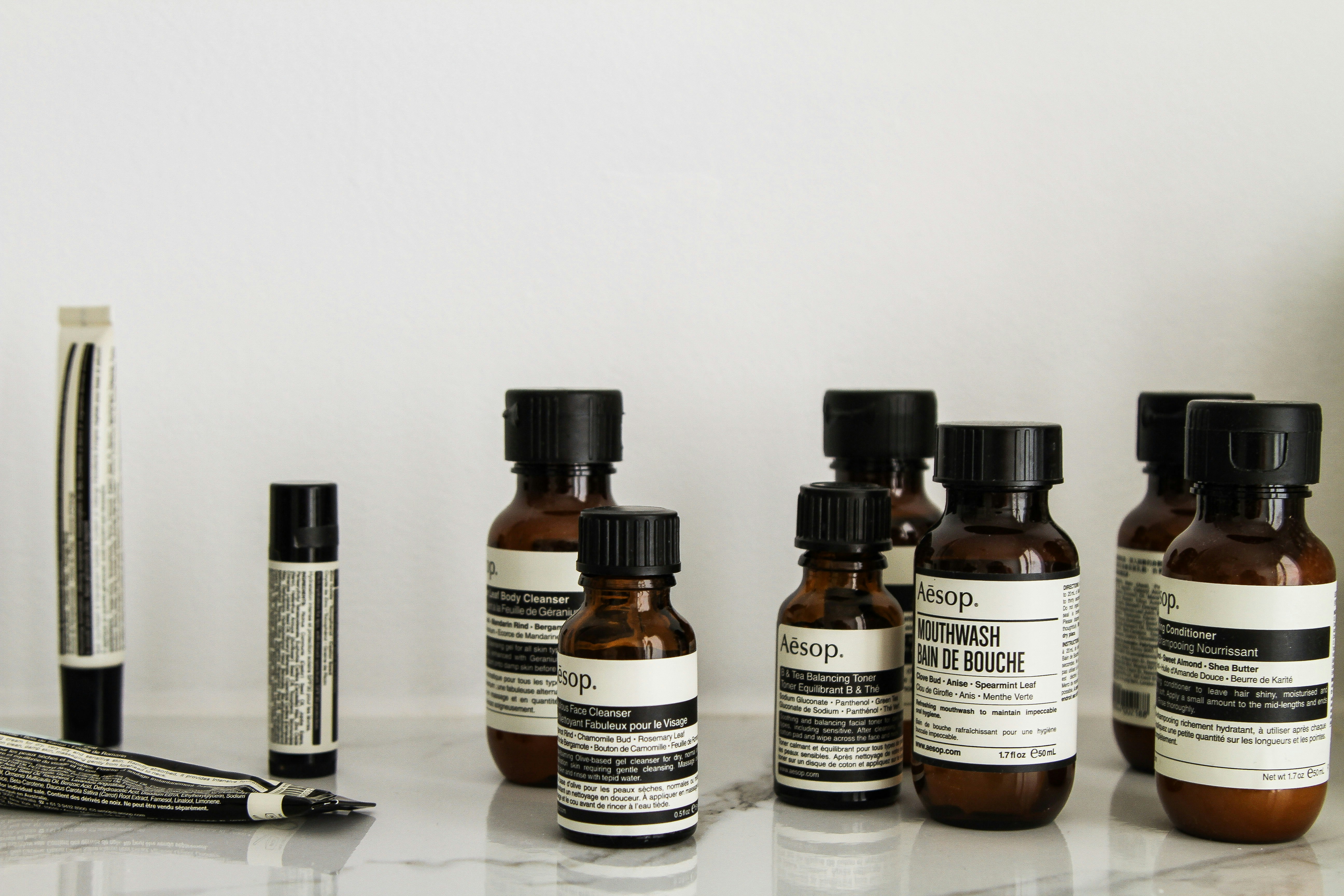 A collection of Aesop branded skincare and grooming products arranged on a light-colored marble surface. The products include small amber glass bottles with black screw caps, a tube, and stick-shaped containers. All labels are black and white, featuring product information in both English and possibly French.