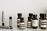A collection of Aesop branded skincare and grooming products arranged on a light-colored marble surface. The products include small amber glass bottles with black screw caps, a tube, and stick-shaped containers. All labels are black and white, featuring product information in both English and possibly French.