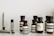 A collection of Aesop branded skincare and grooming products arranged on a light-colored marble surface. The products include small amber glass bottles with black screw caps, a tube, and stick-shaped containers. All labels are black and white, featuring product information in both English and possibly French.