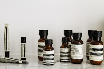A collection of Aesop branded skincare and grooming products arranged on a light-colored marble surface. The products include small amber glass bottles with black screw caps, a tube, and stick-shaped containers. All labels are black and white, featuring product information in both English and possibly French.