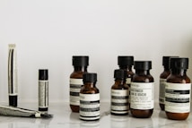 A collection of Aesop branded skincare and grooming products arranged on a light-colored marble surface. The products include small amber glass bottles with black screw caps, a tube, and stick-shaped containers. All labels are black and white, featuring product information in both English and possibly French.