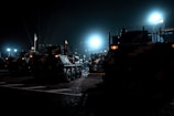 Several military vehicles are positioned in a formation at night under bright artificial lights. Flags are attached to some of the vehicles, and there is an evident presence of military or security personnel preparing or inspecting the vehicles. The overall environment is dark, with the prominent lighting creating stark contrasts and illuminating the scene.