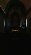 A dimly lit church interior containing an altar with religious iconography, statues, and an intricately designed backdrop. The central focus is on the altar, illuminated with a subtle yellow light that contrasts with the surrounding darkness, creating a serene and contemplative atmosphere.