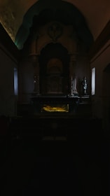 A dimly lit church interior containing an altar with religious iconography, statues, and an intricately designed backdrop. The central focus is on the altar, illuminated with a subtle yellow light that contrasts with the surrounding darkness, creating a serene and contemplative atmosphere.