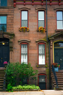 A classic brownstone building facade features three tall windows with decorative framing. Two windows have flower boxes with bright red and yellow flowers. Black metal railings adorn the small garden area, which is filled with lush green plants. Stairs on either side lead to dark-colored doors adorned with flowers in pots.
