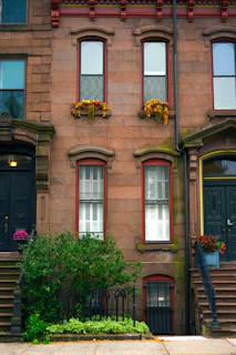 A classic brownstone building facade features three tall windows with decorative framing. Two windows have flower boxes with bright red and yellow flowers. Black metal railings adorn the small garden area, which is filled with lush green plants. Stairs on either side lead to dark-colored doors adorned with flowers in pots.