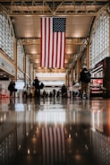 A large American flag hangs from the ceiling in a spacious airport terminal. Floors reflect the lights and flag above, while travelers walk with luggage or wait in groups.