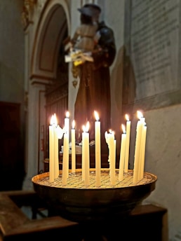 A group of lit candles stands on a circular holder with a grid pattern. Behind the candles, there is a statue in a dimly lit room, likely a church or chapel. The scene conveys a peaceful or devotional atmosphere.