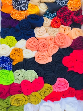 A collection of vibrant, rolled fabrics arranged in a grid pattern, showcasing a variety of colors such as black, red, orange, yellow, blue, and green. Each roll is neatly organized, creating an aesthetically pleasing and colorful display.