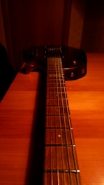 A dark-toned electric guitar lies on a wooden table, with the neck and strings in clear focus while the body fades into the shadows. The warm lighting casts an orange hue across the surface.