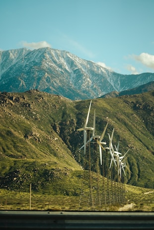 A row of wind turbines is set against a backdrop of a large, snow-capped mountain range. The foreground consists of green rolling hills and a blue sky with some clouds.