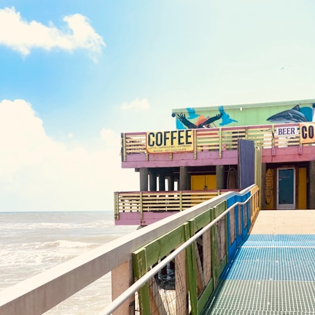 A vibrant, colorful building on a pier with signs advertising coffee and beer. The structure is elevated on pillars over a beach or sea setting. Bright pastel colors are used, and there are various artistic elements on the walls, including a mural with oceanic themes.