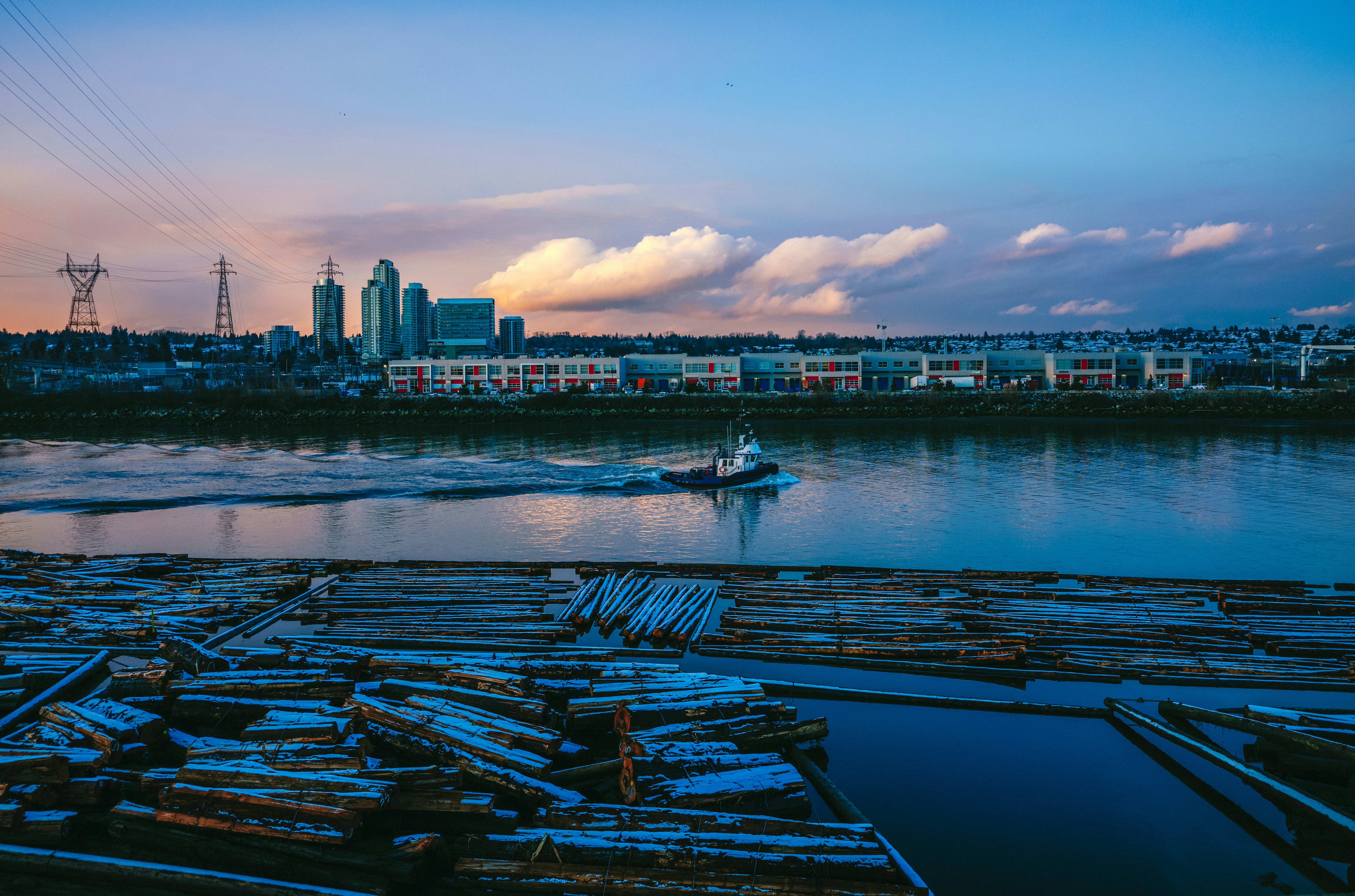 Dusk light over a river with a distant city skyline and stacked logs in the foreground, emphasizing a calm, industrial waterfront scene.